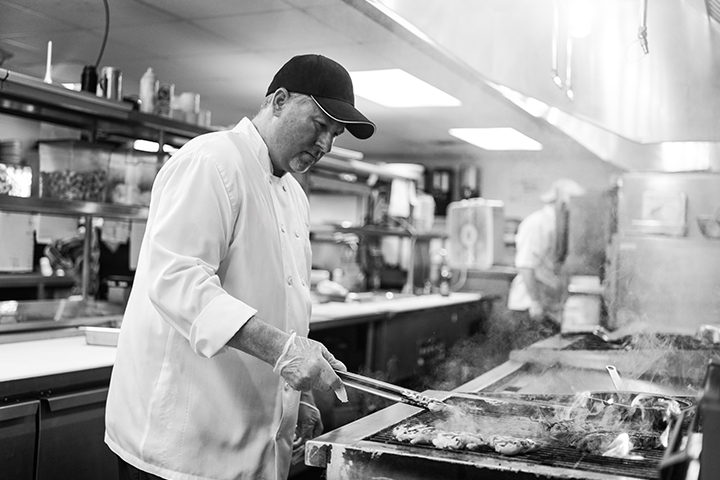 Chef grilling patties in a professional kitchen, wearing a cap and gloves.