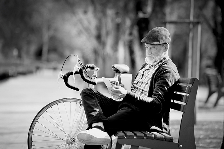 Older man sitting on a park bench with a coffee, looking at his phone; bicycle leaned beside him. Black and white photo.