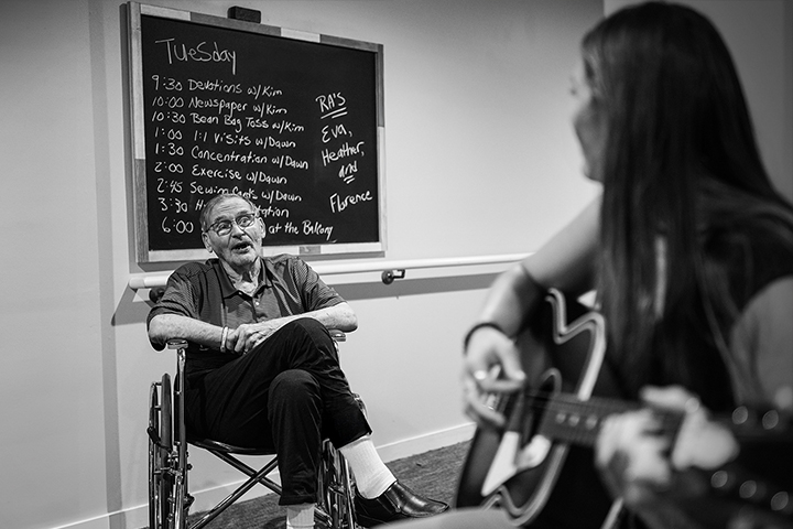 Elderly man in a wheelchair listens to a woman playing guitar, with a daily schedule on a chalkboard behind him. Black and white photo.