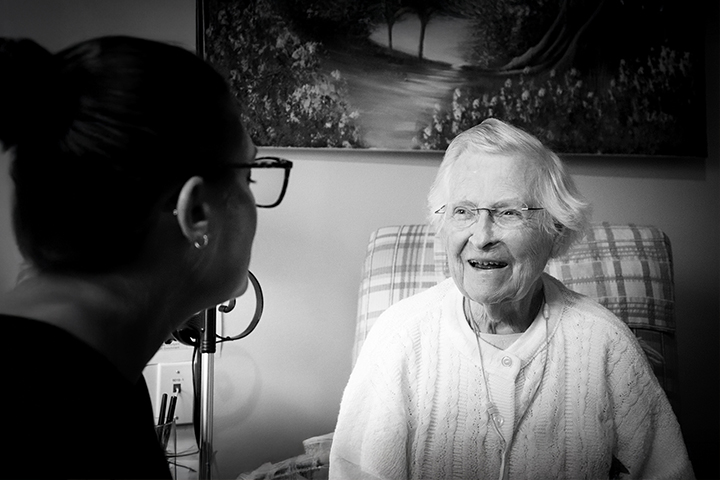 An elderly woman smiles while talking to a caregiver in a cozy, warmly lit room. Black and white photo.