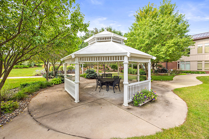 White gazebo with black metal chairs in a landscaped garden near a building on a sunny day.