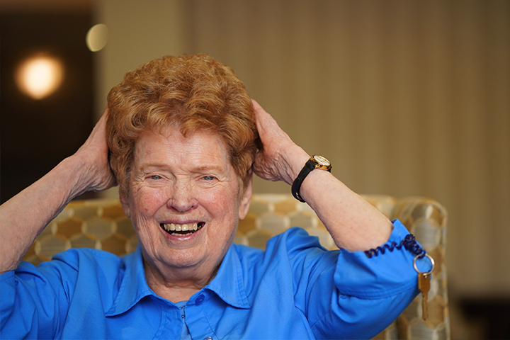 Smiling elderly woman in a blue shirt holds her hands on her head while sitting on a patterned chair.