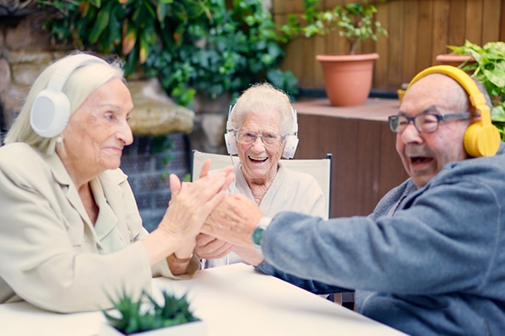 Three elderly people wearing headphones laugh and clap hands together while sitting at a table outdoors.
