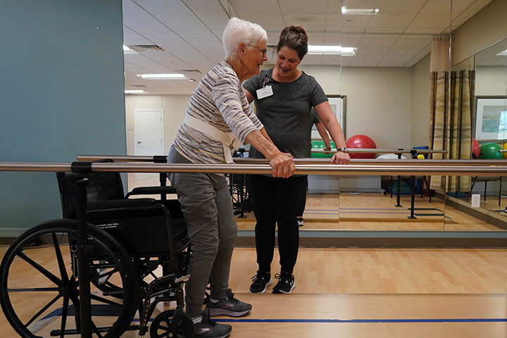An elderly woman in rehab uses parallel bars to walk with assistance from a physical therapist.
