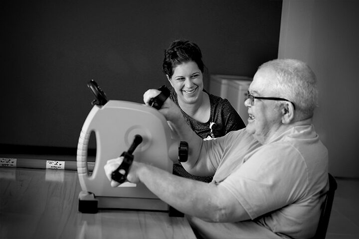 An older man exercises with a hand bike while a woman beside him smiles supportively.