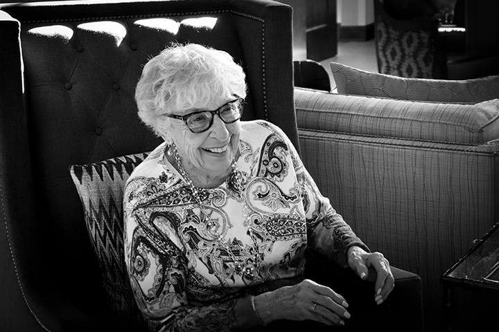 Elderly woman with glasses smiling while sitting in an armchair, wearing a patterned top.