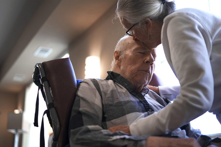 An elderly woman kisses the forehead of an elderly man in a wheelchair, showing tenderness and care.