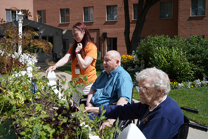 Three people sit near a garden outside, two elderly in wheelchairs and one woman standing, enjoying the sunny day.