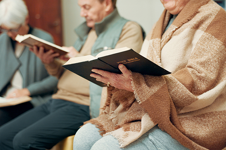 Three elderly people sitting and reading Bibles, one wrapped in a brown blanket.