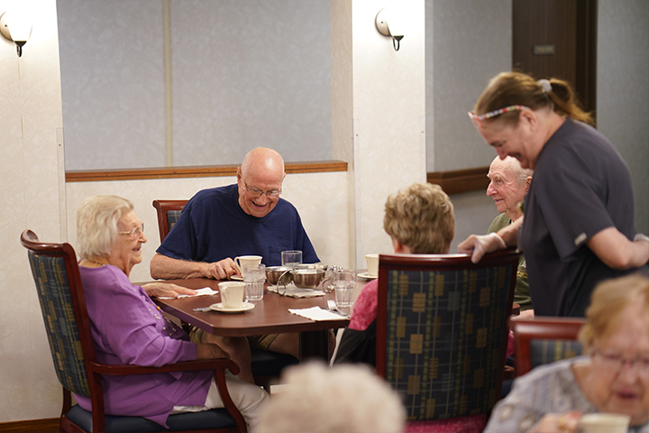 Four elderly people sit smiling at a dining table while a caregiver stands nearby, also smiling.