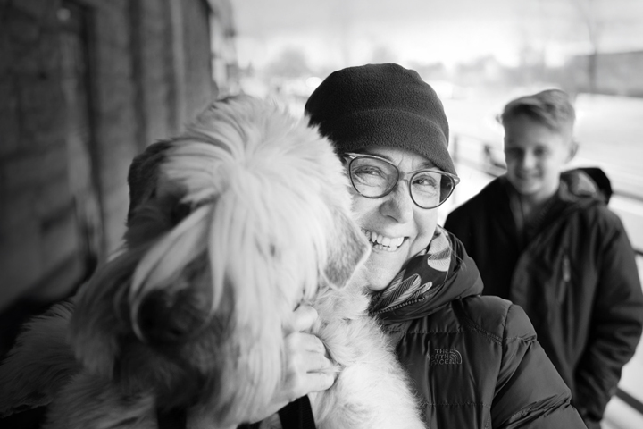 Smiling person holding a fluffy dog, with another person in the background on a snowy day.