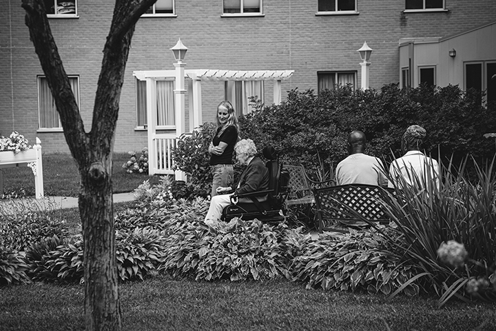 Four elderly people sit smiling at a dining table while a caregiver stands nearby, also smiling.