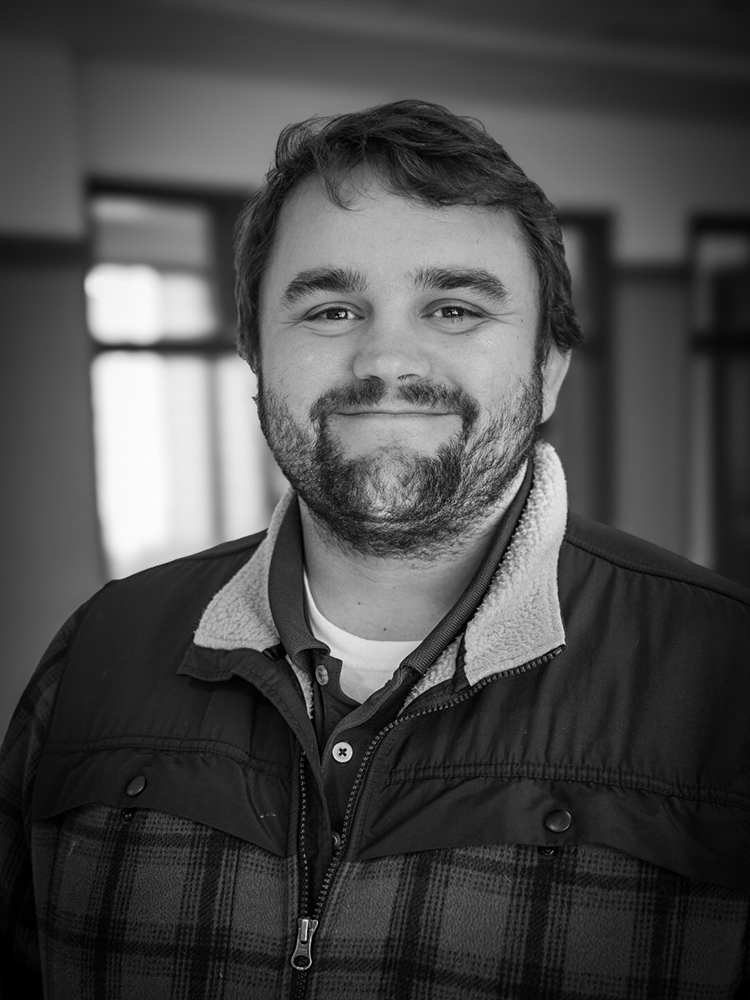 John Linn, a bearded man, smiles at the camera in a plaid jacket indoors, captured in black and white.