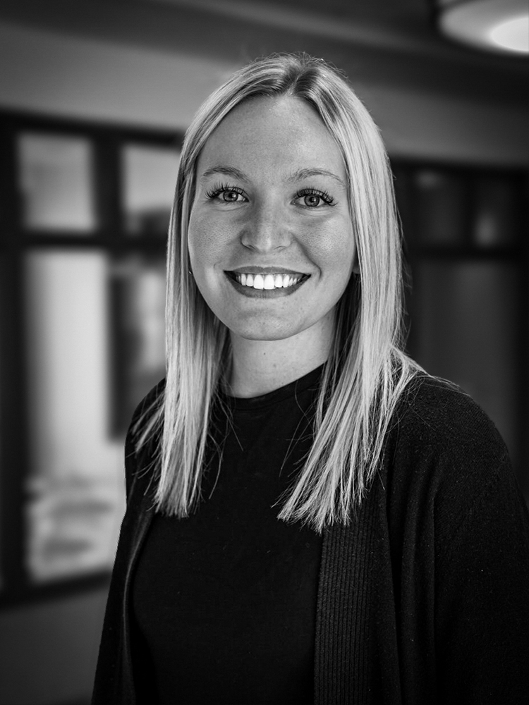 Hailey Richter, a smiling woman with straight, blonde hair wearing a dark top, stands indoors with blurred background.