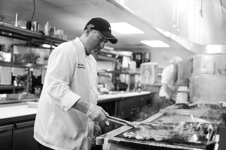 Chef in a white coat and cap grilling meat in a busy, professional kitchen.