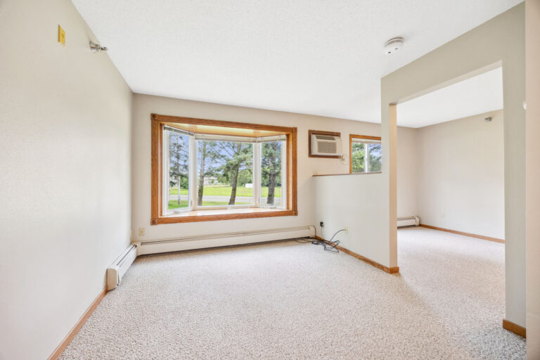 Empty assisted living room with a large bay window, white walls, and beige carpet, offering a view of green trees outside.