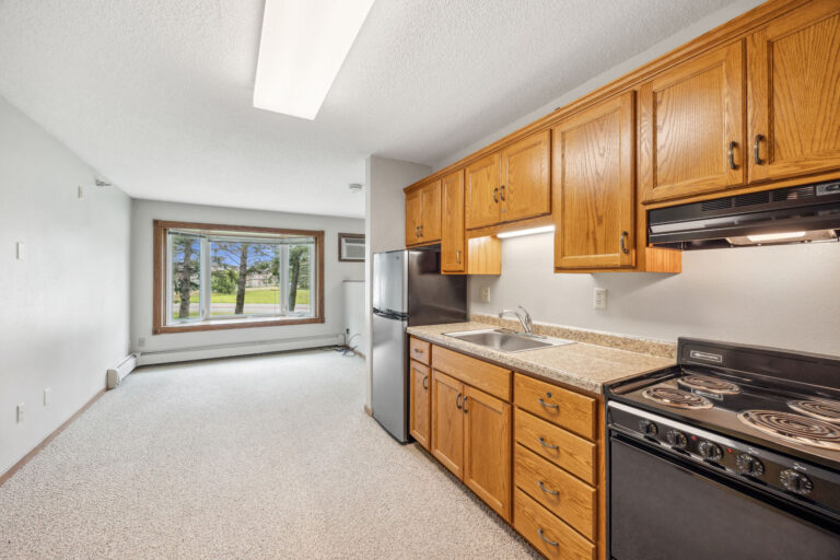 Compact Benedict Court apartment kitchen with wooden cabinets and appliances, next to a living area with a large window.