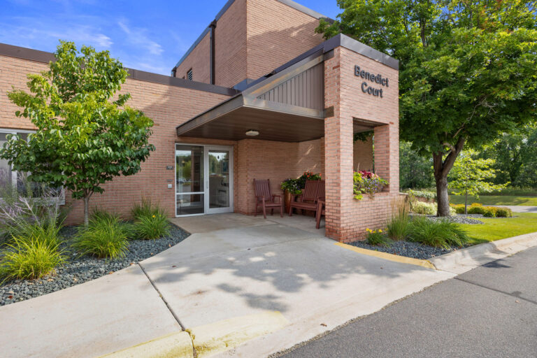 Benedict Courts East Entrance features a brick facade, lush plants, and cozy chairs by the door.