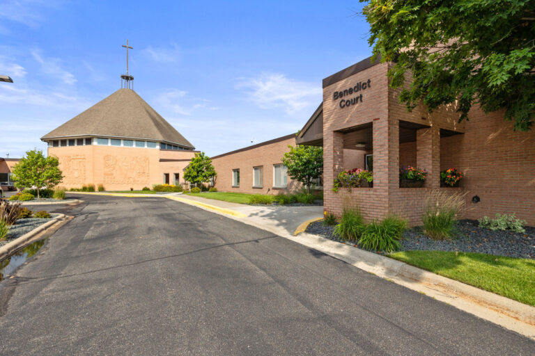 Conical-roofed chapel at Benedict Court East Entrance, with a sign, lush greenery, and charming flower boxes.