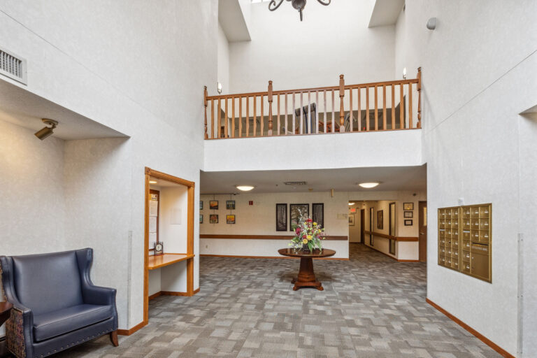 The Benedict Court entrance boasts a spacious lobby with a high ceiling, floral table, mailboxes, blue chair, and wooden railing.