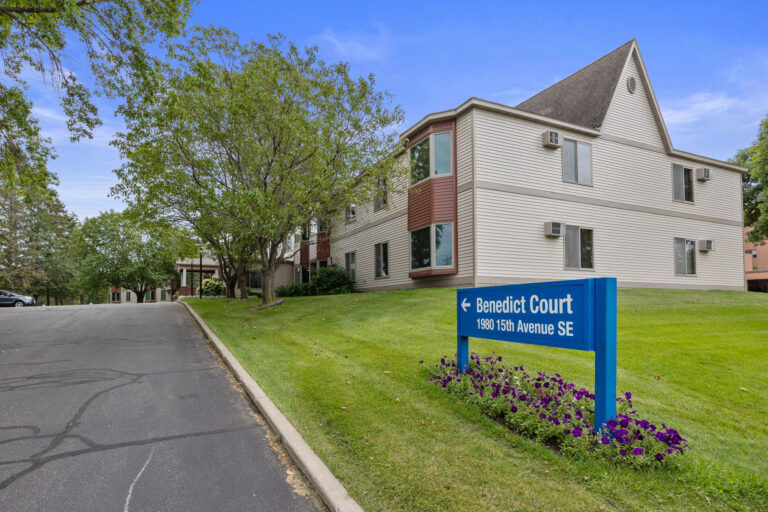 Two-story building with sign reading Benedict Court, surrounded by trees and grass, on a clear day.
