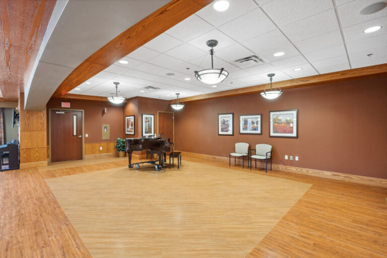 The Bonn Great Room features wooden floors, brown walls, a piano, chairs, framed artworks, and is beautifully lit by ceiling lights.