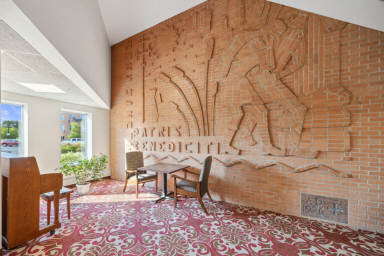 The Chapel Dedication Wall showing in brick relief art, patterned carpet, two chairs, and large windows showing outdoor greenery.