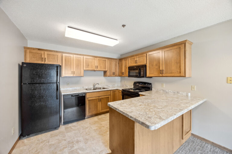 Benedict Village Apartment kitchen with wooden cabinets, black appliances, and a marble-pattern countertop under overhead light.