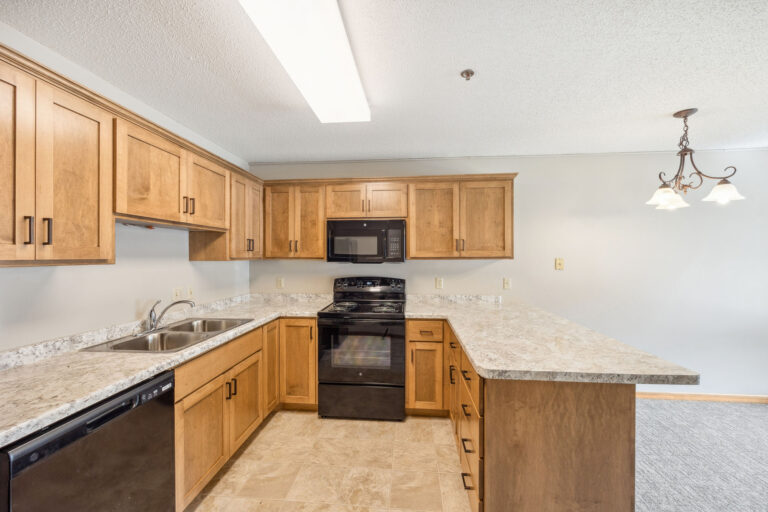 Benedict Village Apartment kitchen with wooden cabinets, black appliances, and a marble-pattern countertop under overhead light.