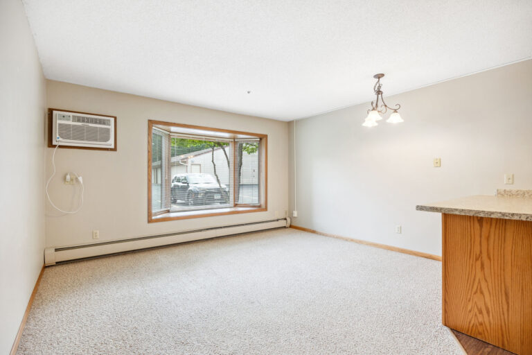 Cozy living room at Benedict Village, featuring a large window, AC unit, beige carpet, and a light near the kitchen counter.