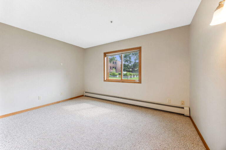 Benedict Village bedroom with beige walls, carpet, and garden-view window. Ceiling light warmly lit.