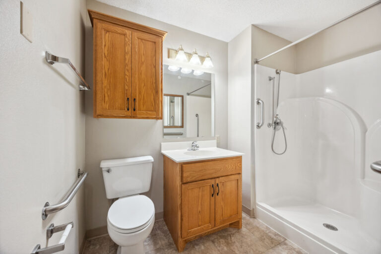 Benedict Village Apartment Bathroom: Wooden cabinets, sink, toilet, and walk-in shower on white walls and tile floor.
