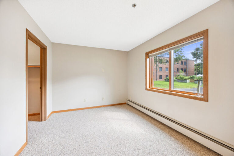 Bedroom in Benedict Village with beige carpet, white walls, and a wooden-trimmed window overlooking trees and a brick building.