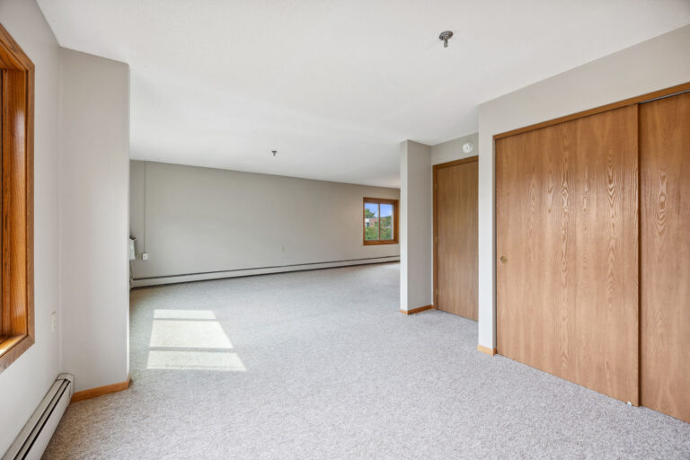 Benedict Village Apartment living room with beige carpet, wooden closets, light walls, and a window letting in natural light.