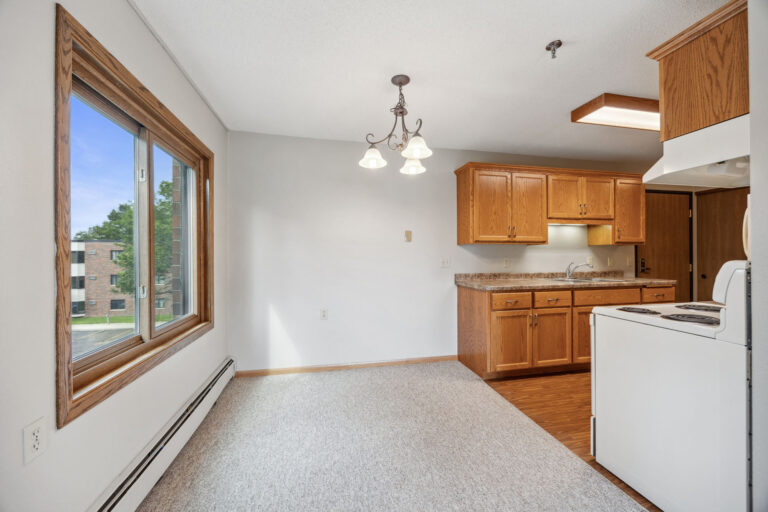 Dining area at Benedict Village Apartment with wooden cabinets, white walls, large window, and overhead light fixture.