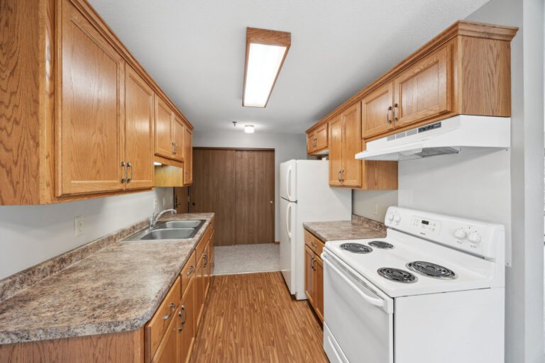 Benedict Village Apartment kitchen: a narrow space with wooden cabinets, white appliances, and marble-patterned countertops.