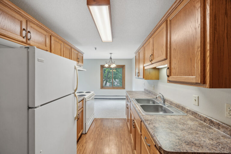 Benedict Village Apartment kitchen: a narrow space with wooden cabinets, white appliances, and marble-patterned countertops.