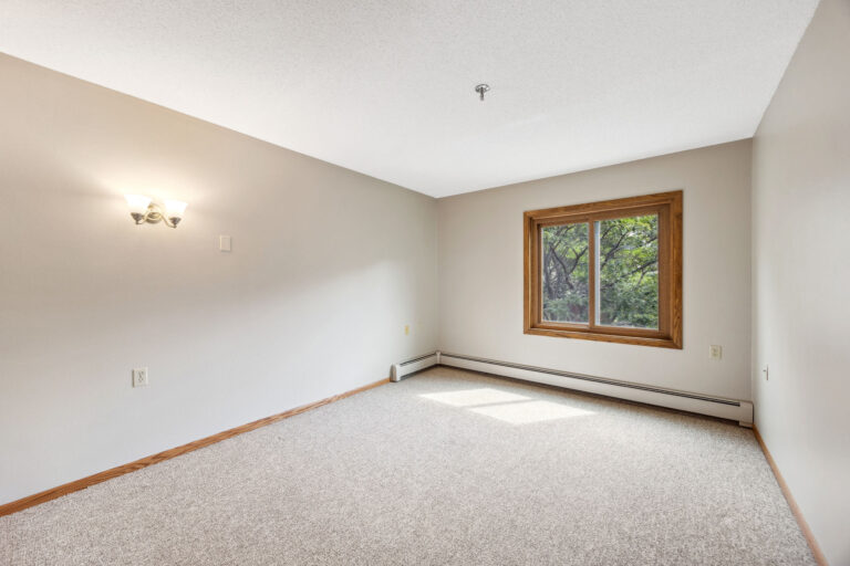 Benedict Village Apartment bedroom: beige room with carpet, a window with wooden trim, and a wall sconce.