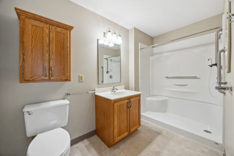 Benedict Village Apartment bathroom: Wooden vanity, mirror, toilet, and white shower-tub combo beneath a ceiling light.