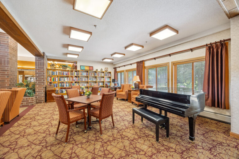 The Benedict Village Library Lounge boasts a cozy space with a grand piano, bookshelves, and warm-lit tables and chairs.