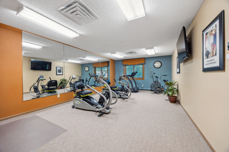 Benedict Village Fitness Area: Cozy gym room with exercise bikes, wall mirrors, a TV, and a clock on orange and blue walls.