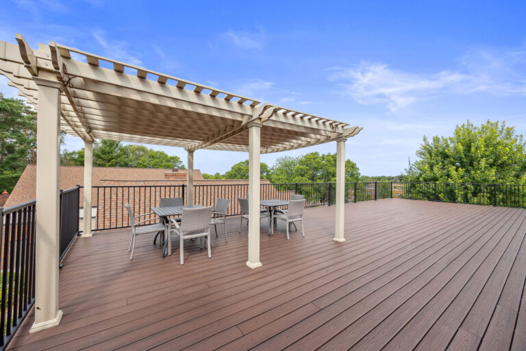 Benedict Village Rooftop Terrace boasts a pergola, table, and chairs, all framed by a black railing under a clear blue sky.