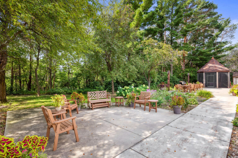 Patio with wooden chairs and benches at Benedict Village Gardens, surrounded by lush greenery, leading to a small gazebo.