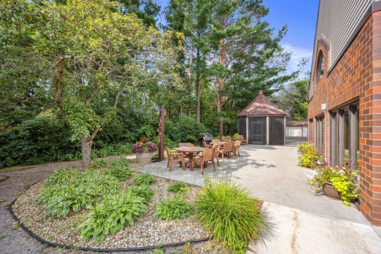 Patio with wooden furniture, a grill, and gazebo in Benedict Village Gardens, surrounded by trees beside a brick building.
