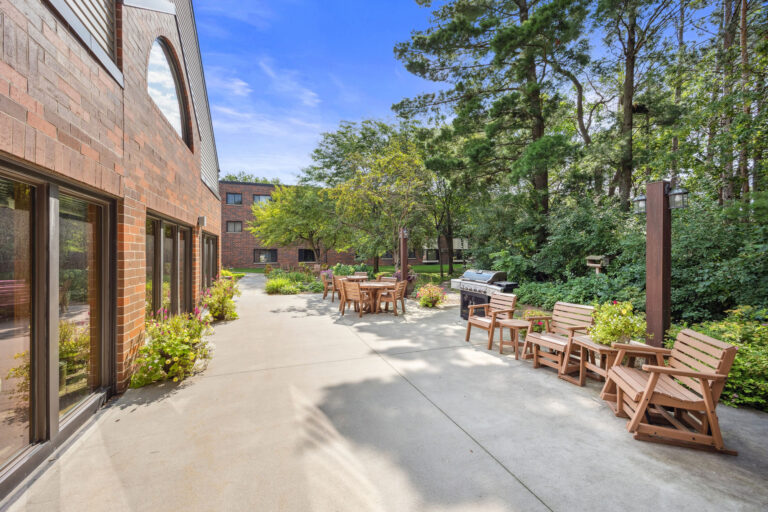 Benedict Village patio and grill area with wooden chairs, surrounded by greenery and brick buildings under a blue sky.
