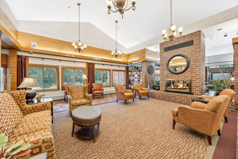 Cozy fireplace lounge in Benedict Village with armchairs, patterned sofas, chandeliers, and a brick fireplace under a high ceiling.