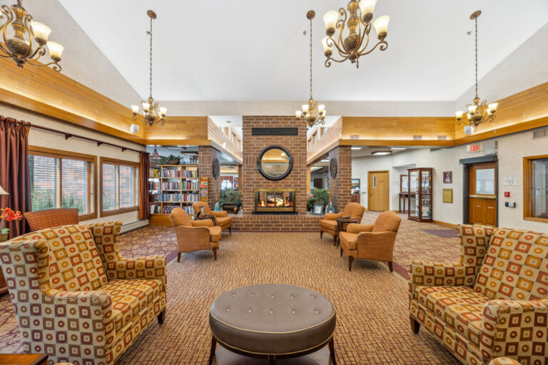 Cozy fireplace lounge in Benedict Village with armchairs, patterned sofas, chandeliers, and a brick fireplace under a high ceiling.