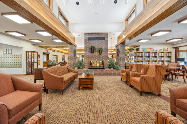 Spacious living room with brown sofas, a central brick fireplace, large windows, and bookshelves reminiscent of Benedict Village Library Lounge.