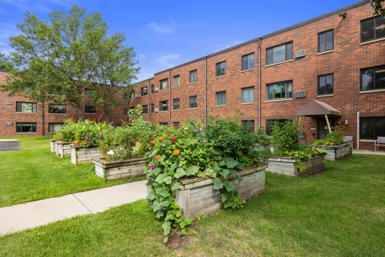 Community garden with vegetables and flowers in raised beds outside the brick Benedict Village apartment building.