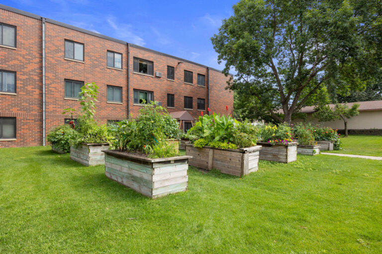 Community garden with vegetables and flowers in raised beds outside the brick Benedict Village apartment building.